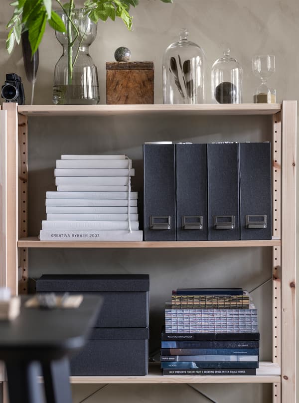 Des étagères en bois montées au mur, sur lesquelles se trouvent des piles de livres et des boîtes de rangement noires, ainsi que des vases et cloches en verre avec des fleurs et des plumes.