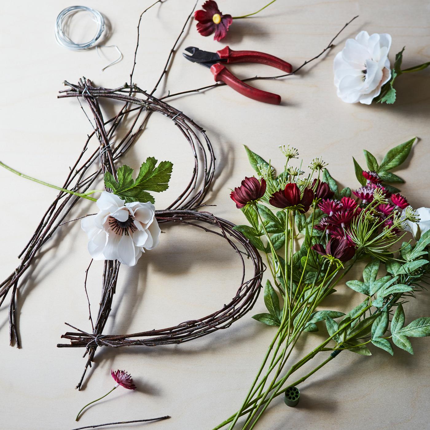 Des fleurs et des branches en pleine transformation pour devenir une décoration florale en forme de B posées sur une surface avec une paire de pinces.