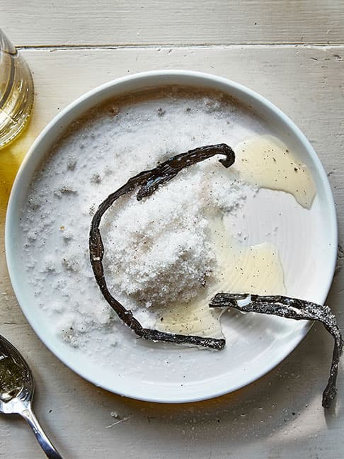 Des gousses de vanille reposent dans un bol de sucre sur une table rustique, mettant en valeur textures et simplicité.