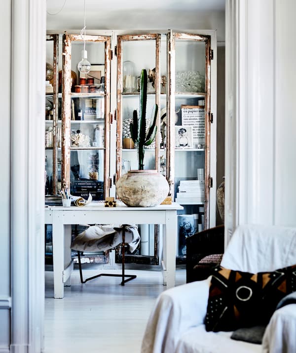 Des objets décoratifs et des bocaux dans une armoire blanche à portes vitrées, derrière un bureau blanc.