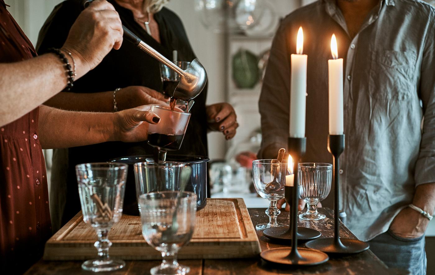 Des personnes autour d’un îlot de cuisine avec des verres et des bougies allumées et une casserole de vin chaud, une personne remplit les verres.