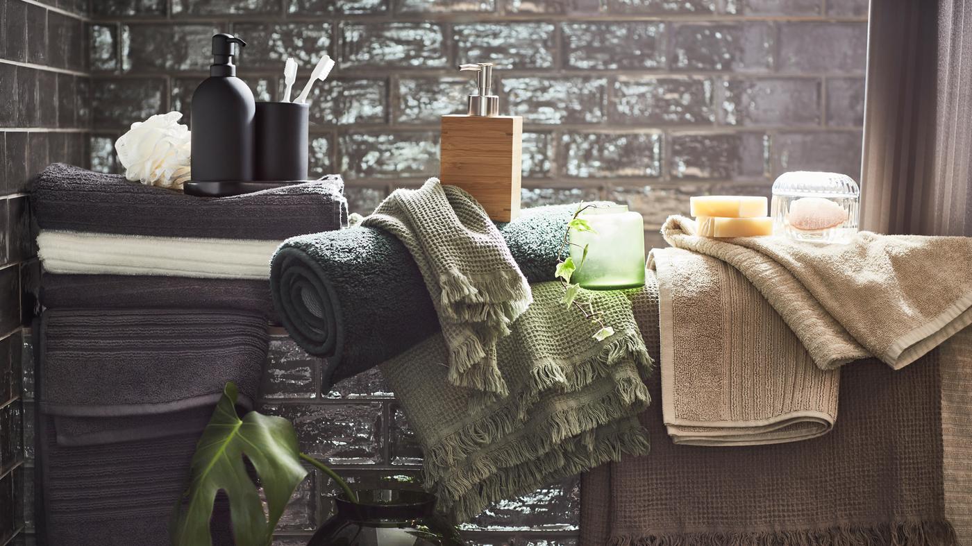 Des serviettes et draps de bain blanches, beige clair, brun/gris clair, gris foncé et vert clair dans une salle de bain aux carreaux gris.