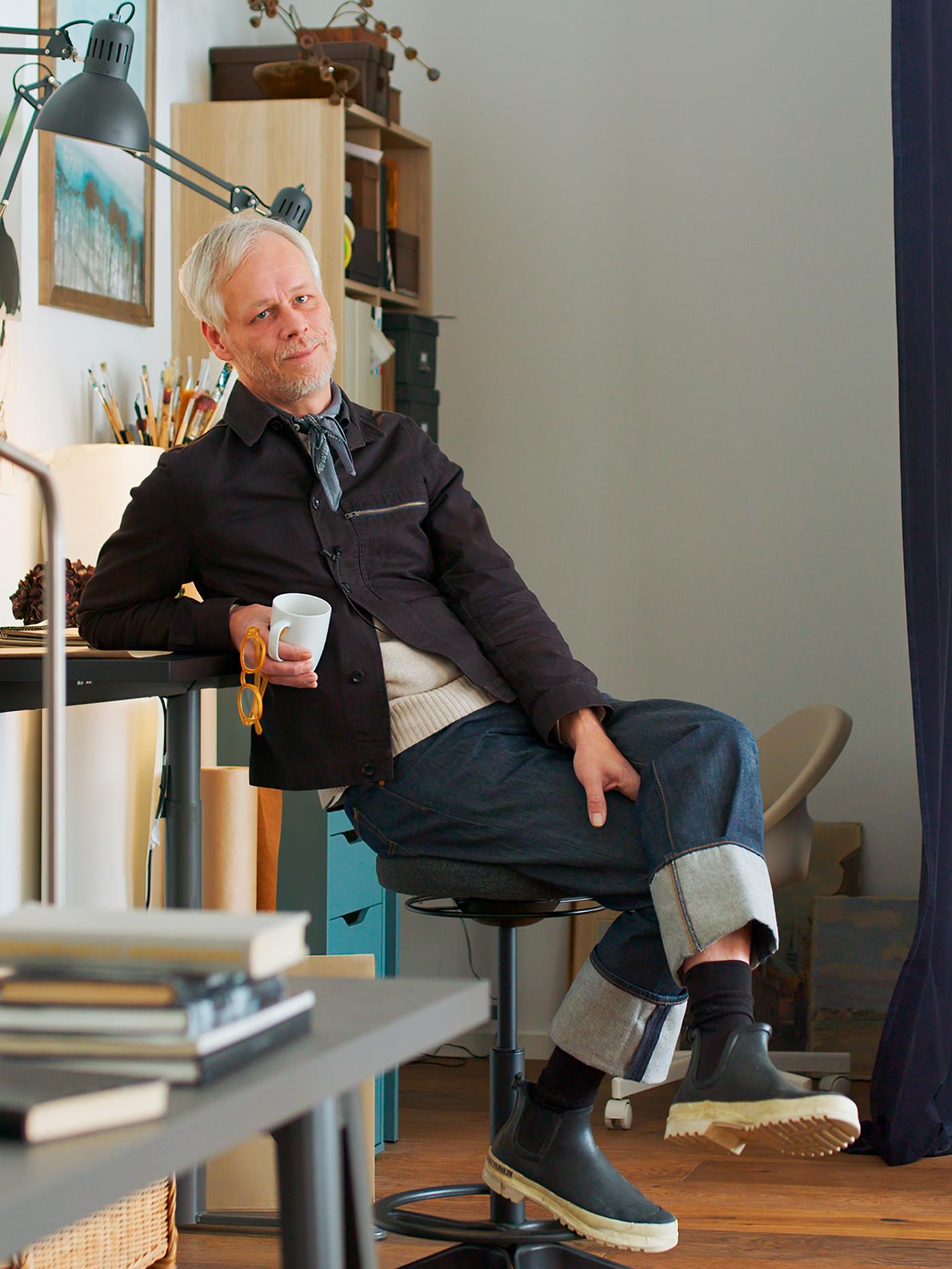 Designer Hans Blomquist sitting on a stool in a light room holding a cup in his hand.