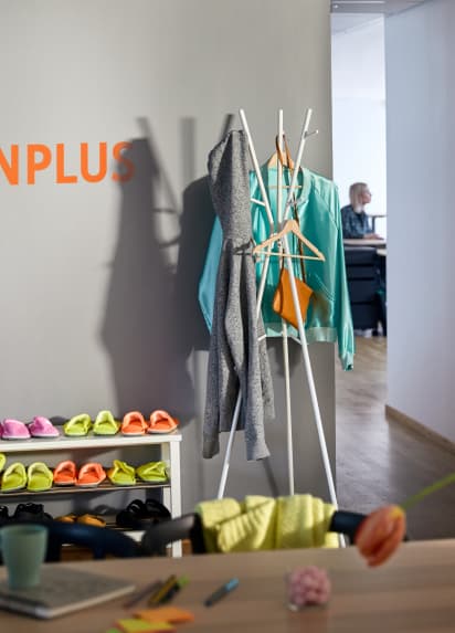 Desk with mug and notepad on it in the foreground with a shoe rack and coat hanger against a wall in the background