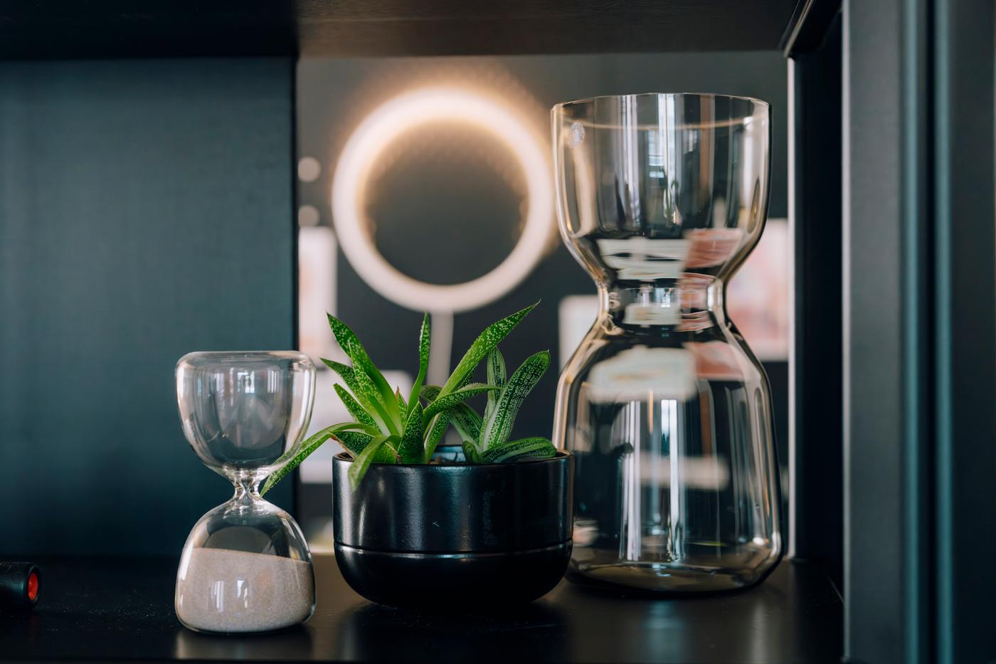 Dettaglio di scaffale con caraffa in vetro, piccola clessidra, vaso nero con pianta e ring light sullo sfondo.