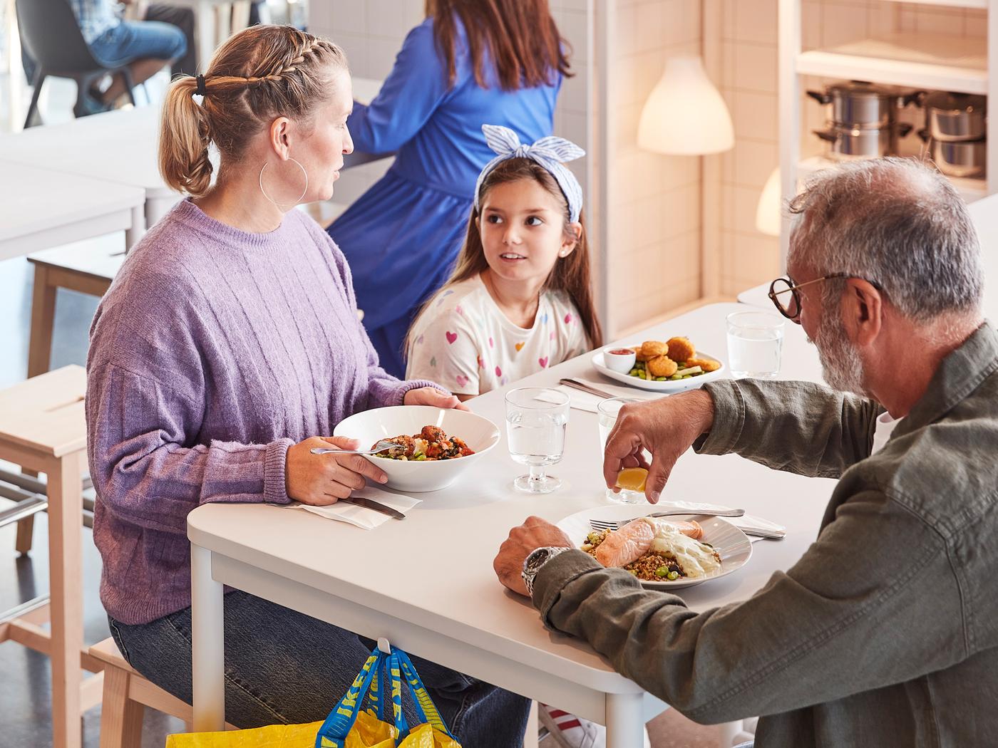 Deux adultes et un enfant sur le point de déguster un repas à la table blanche d’un restaurant IKEA.