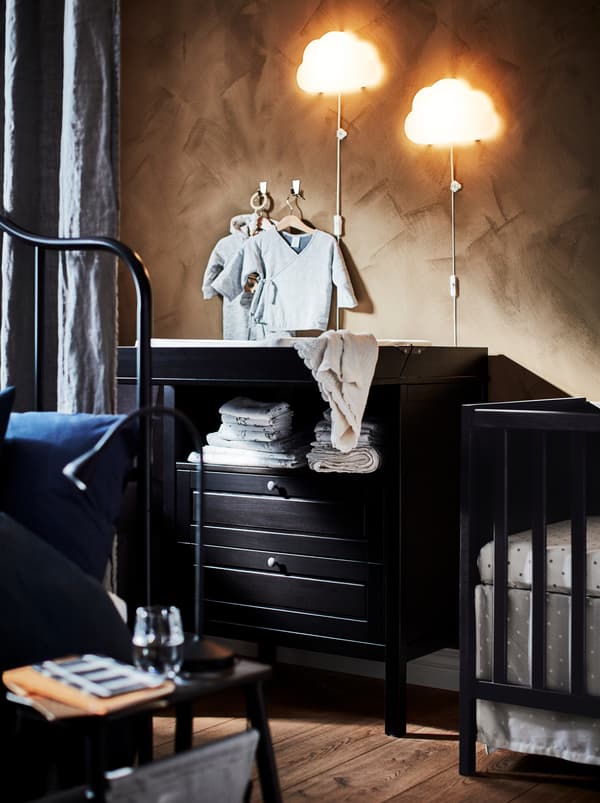 Deux appliques à DEL UPPLYST en forme de nuage sont installées au-dessus d’une table à langer noire SUNDVIK en bois à deux tiroirs se trouvant à côté d’un lit à barreaux.