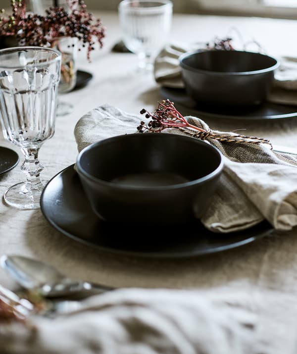 Deux couverts dressés sur une nappe en lin, chacun composé d’un bol gris-bleu sur une assiette avec un verre et une serviette nouée.
