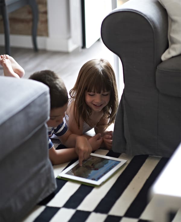 Deux enfants à plat ventre par terre entre deux canapés, en train de jouer avec une tablette à écran tactile.