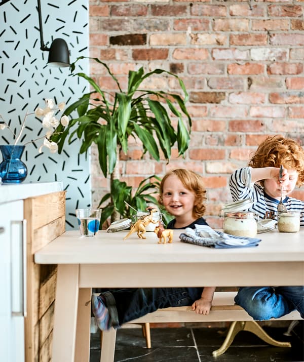 Deux enfants assis à une table en bois clair sur laquelle se trouve le petit déjeuner et des jouets, devant un mur de briques rouges.