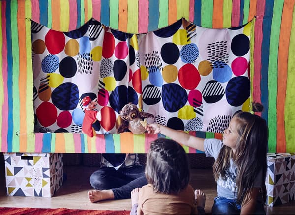 Deux enfants en train de regarder un vieux hibou dans le théâtre de marionnettes pendant qu’un troisième enfant est assis en tailleur derrière le rideau.