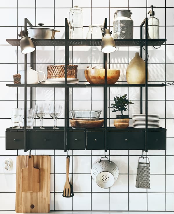 Deux étagères métalliques noires avec assiettes, verres et ustensiles de cuisine, sur un mur carrelé de blanc.