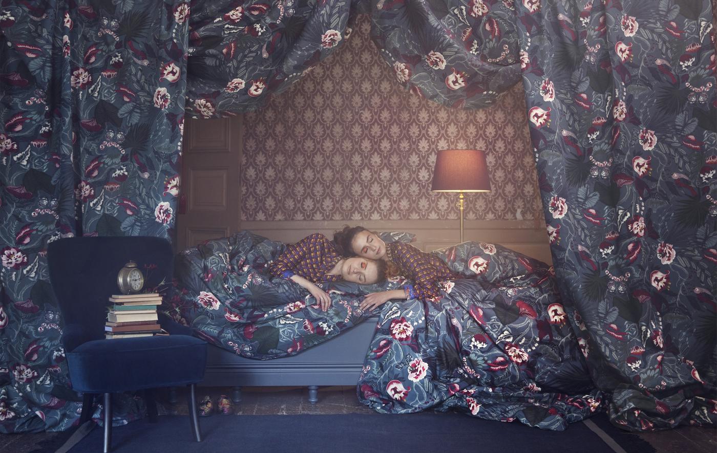 Deux femmes allongées sur un lit, avec papier peint au mur encadré d’un tissu à motif floral.