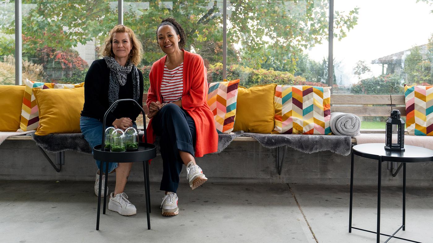 Deux femmes sont assises sur un banc paré de coussins colorés et de fourrures synthétiques. Elles sourient à la caméra