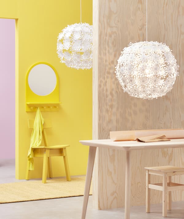 Deux grandes suspensions blanches ornées de motifs floraux, accrochées dans une pièce rose et jaune revêtue de bois clair.
