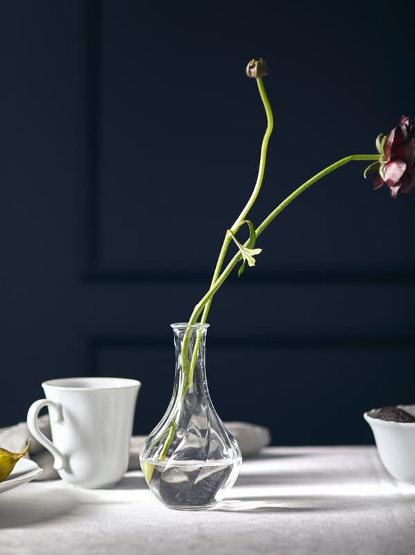 Deux longues tiges de fleur élancées déposées dans un petit vase en verre VILJESTARK court sur une nappe blanche.