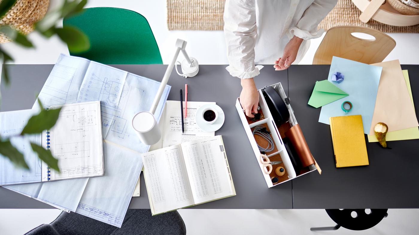 Deux mains prenant un article dans un range-tout de bureau blanc sur deux tables jointes avec des papiers et une lampe de travail blanche.