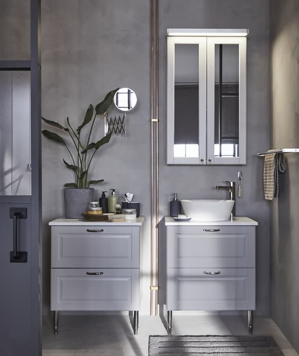 Deux meubles de rangement pour salle de bain gris, l’un avec un lavabo et une armoire avec miroir fixée au-dessus sur le mur gris.