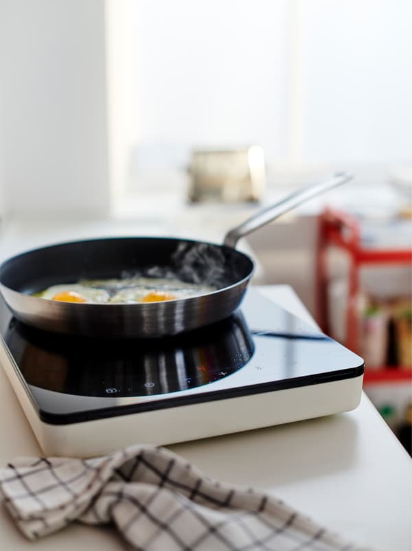 Deux œufs cuisant dans une poêle sur une table de cuisson à induction mobile TILLREDA placée sur un plan de travail, un torchon posé à côté.