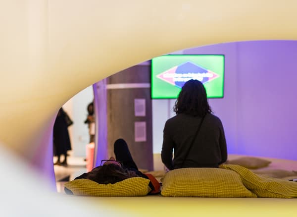 Deux personnes assises sur des coussins regardant un film dans un espace d’exposition.