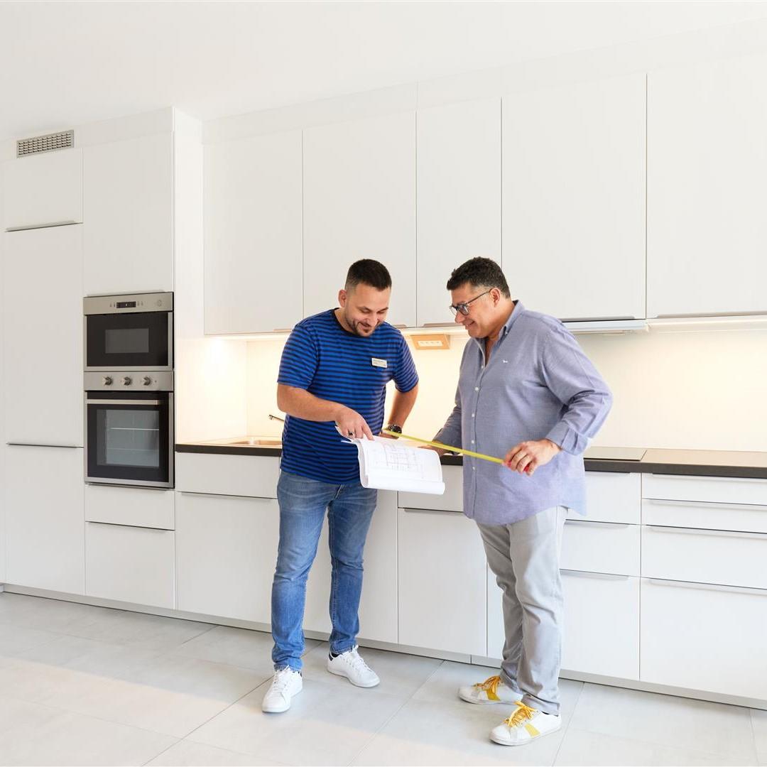 Deux personnes dans une cuisine moderne, avec mètre et clipboard; armoires blanches, four et micro-ondes encastrés.