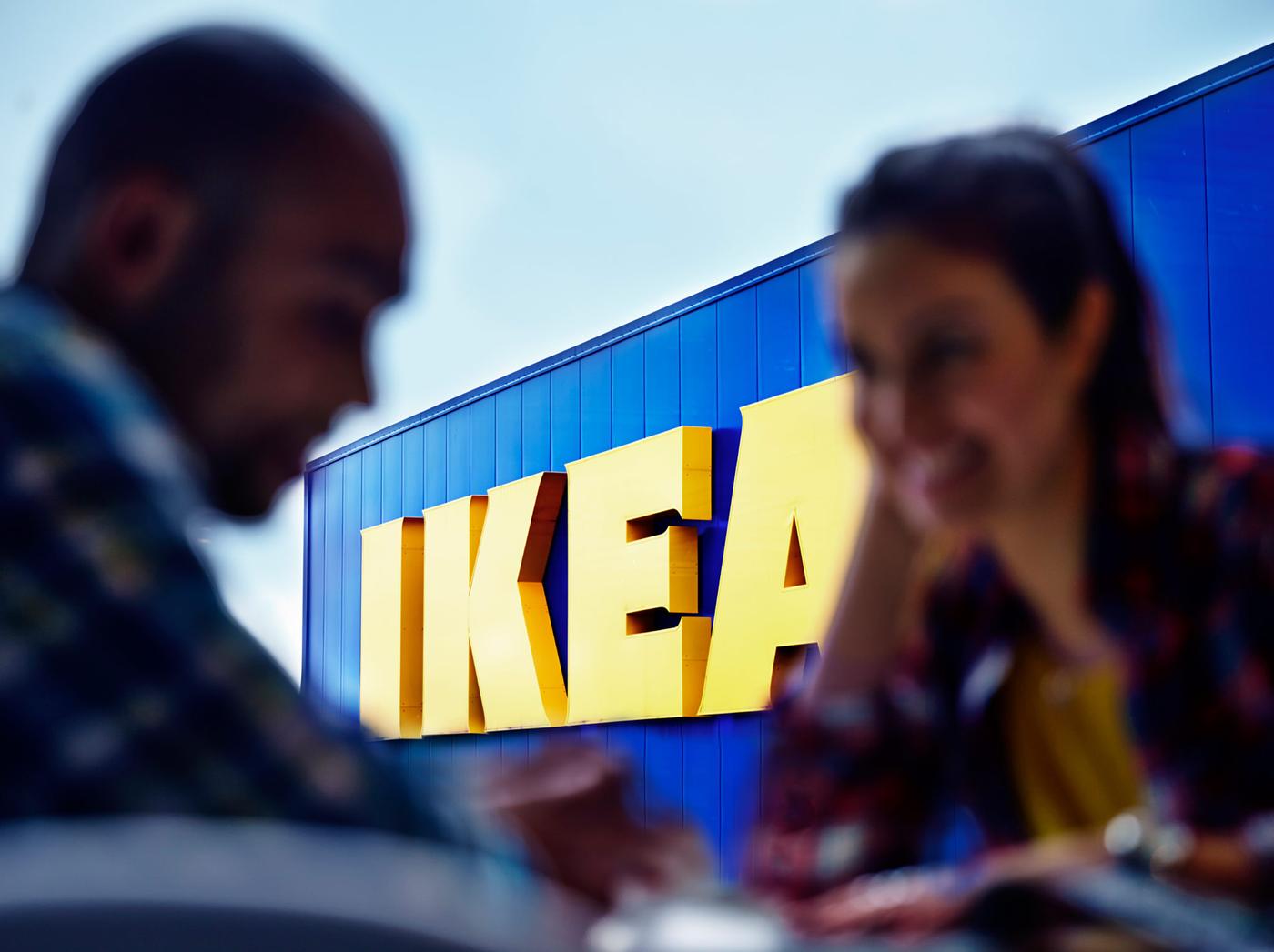 Deux personnes discutent devant le logo d’un magasin IKEA. Le logo jaune sur fond bleu reproduit les couleurs du drapeau de la Suède.