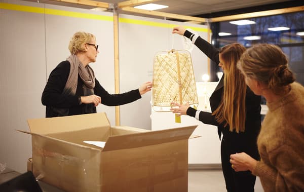 Deux personnes regardent Anna Granath qui tient un luminaire tressé au-dessus d’une grosse boîte.
