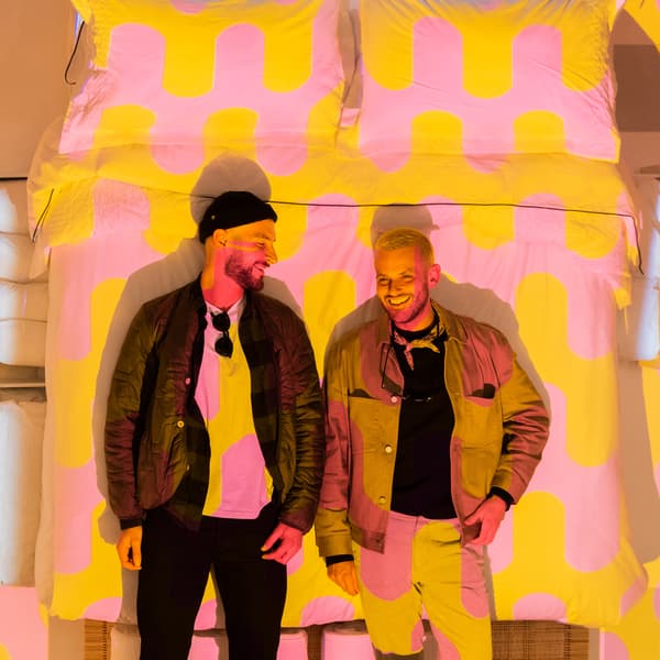 Deux personnes se tiennent devant une image d'un lit garni de linge de lit rose et jaune.