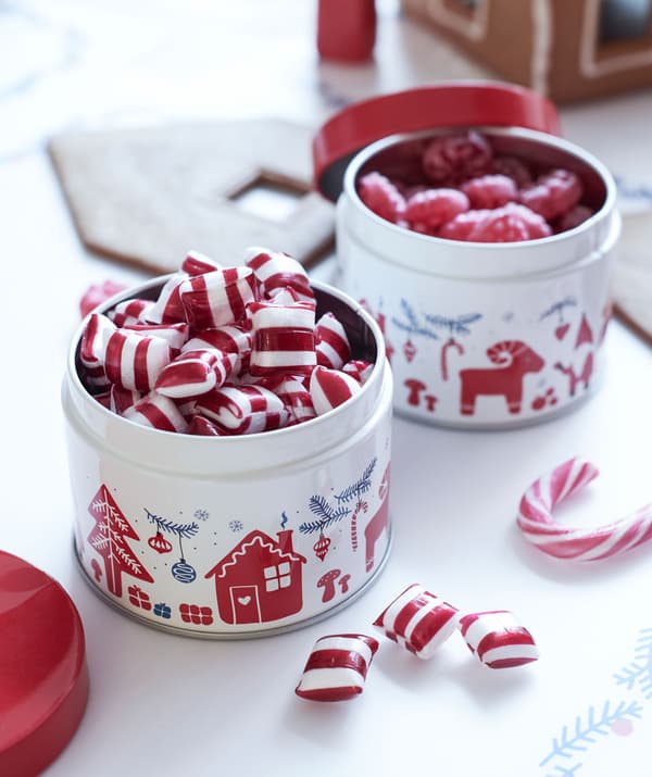 Deux petites boîtes en métal YULETIDE à motifs remplies de bonbons durs, le tout posé sur un comptoir à proximité d’une maison en pain d’épices en construction. 