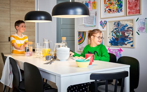 Deux suspensions NYMÅNE noires au-dessus d’une grande table blanche où sont installés deux enfants. Au mur, des œuvres d’enfant encadrées. 