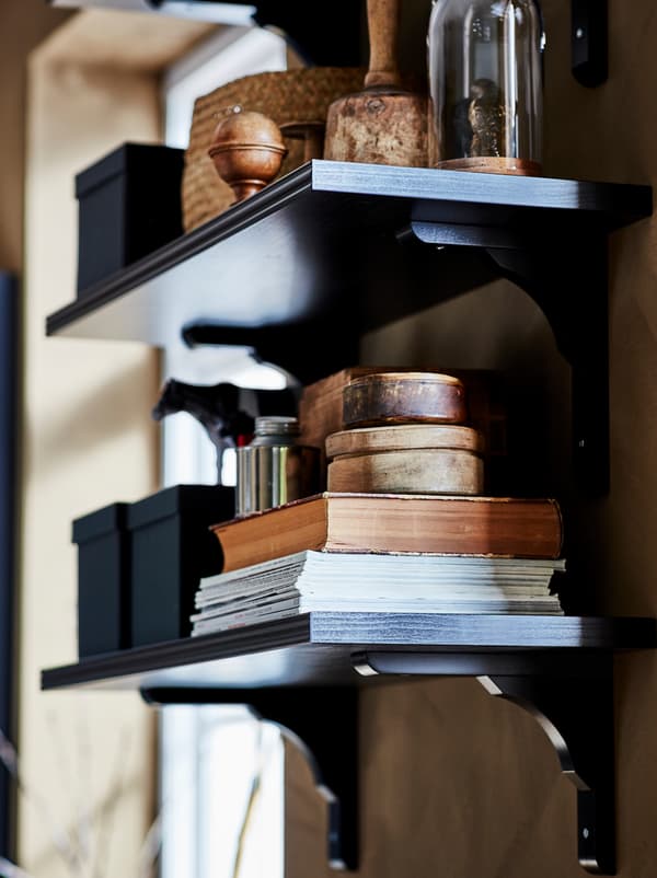 Deux tablettes murales brunes/noires BERGSHULT/RAMSHULT sur lesquelles sont rangés des livres, des boîtes et des objets décoratifs.
