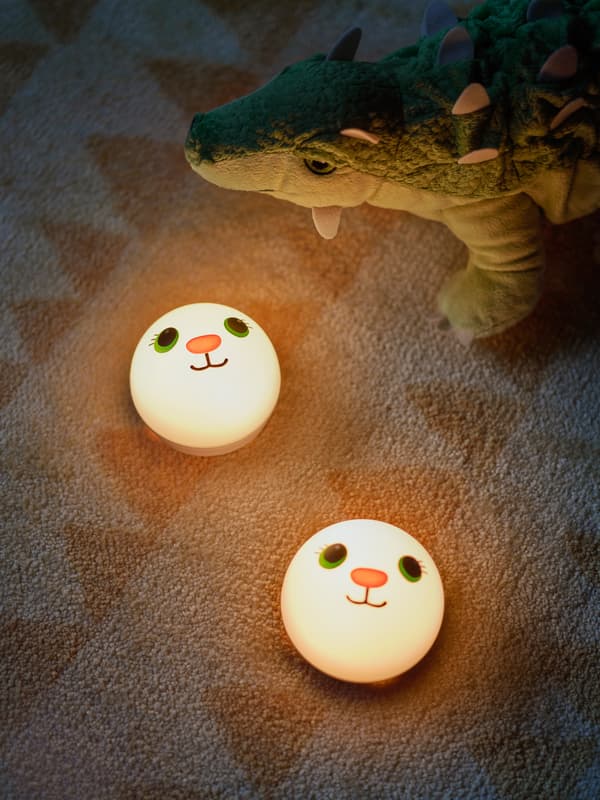 Deux veilleuses KORNSNÖ sont sur un tapis aux motifs colorés pour enfant, alors qu’un dinosaure JÄTTELIK se tient derrière.