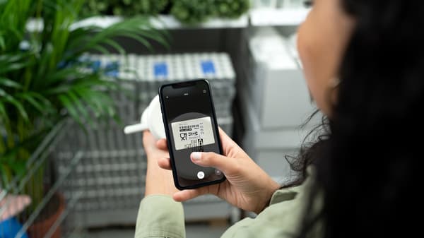 Devojka se nalazi u robnoj kući IKEA. U jednoj ruci drži telefon u drugoj telefon. Na telefonu je otvorena IKEA aplikacija i IKEA Scan opcija. Ona skenira proizvod.