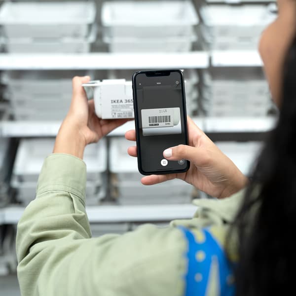 Devojka se nalazi u robnoj kući IKEA. U jednoj ruci drži telefon u drugoj telefon. Na telefonu je otvorena IKEA aplikacija i IKEA Scan opcija. Ona skenira proizvod.