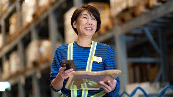 倉庫で商品を確認するスタッフ。青いユニフォームと安全ベストを着用し、棚に並ぶ商品が背景。