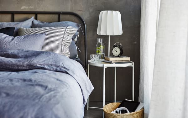 Die Seite eines gemachten Bettes mit Ablagetisch, darauf u. a. IKEA 365+ Karaffe mit Stöpsel aus Klarglas/Kork, Bücher, ein Wecker und eine Leuchte.