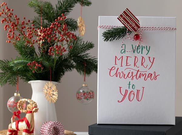 Die TJENA Schachtel wird mit Geschenkanhängern, Geschenkbändchen und Dekokugeln dekoriert.