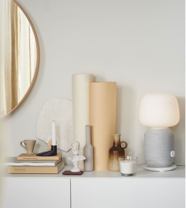 Différents objets de décoration placés sur une surface blanche, avec notamment une lampe-enceinte SYMFONISK grise et blanche.