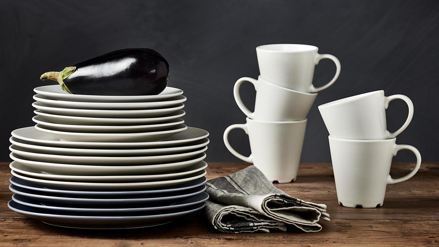 DINERA beige/white mugs are stacked on a worktop next to a stack of beige and grey/blue plates in various sizes.