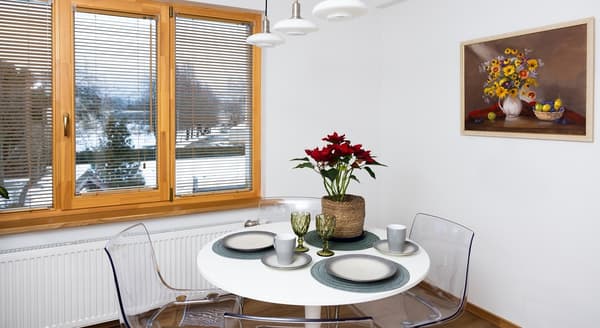 Dining area with round white table, transparent chairs, and red poinsettia centerpiece by a large window.