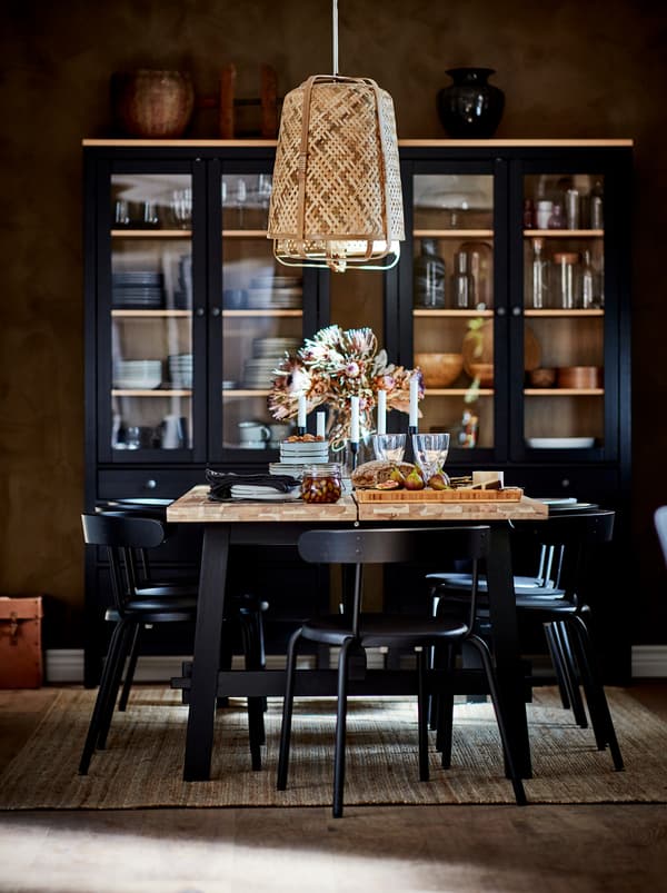Dining table being festively set under a KNIXHULT pendant. Dark glass-door cabinets with table accessories by the far wall.