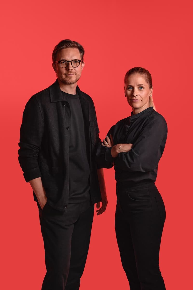 Diseñadores S Fager y H Preutz vestidos compleamente de negros y atrás un fondo de color rojo.