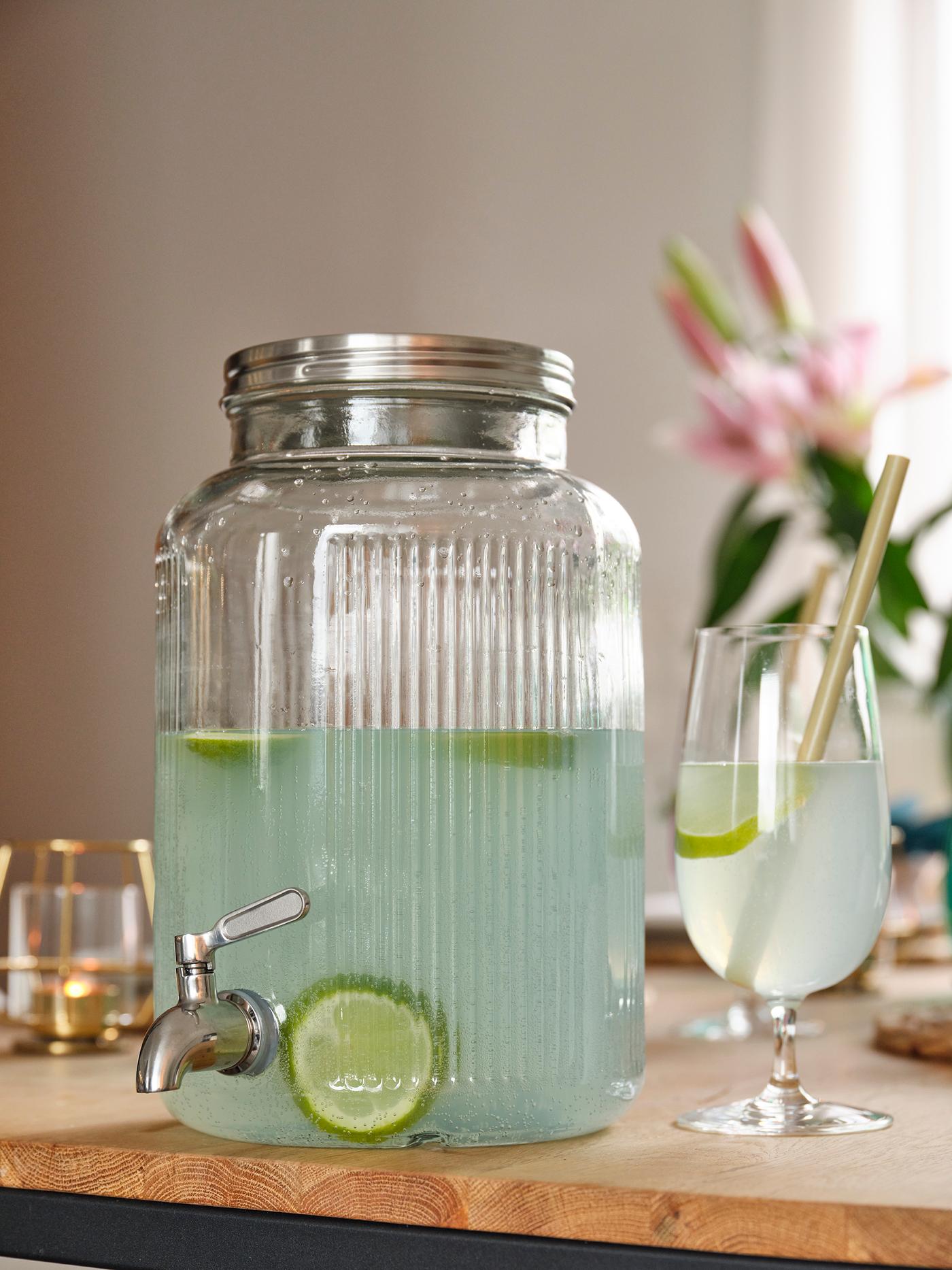 Distributore di bevande in vetro con acqua al lime, rubinetto e bicchiere con cannuccia su una tavola imbandita in stile estivo.