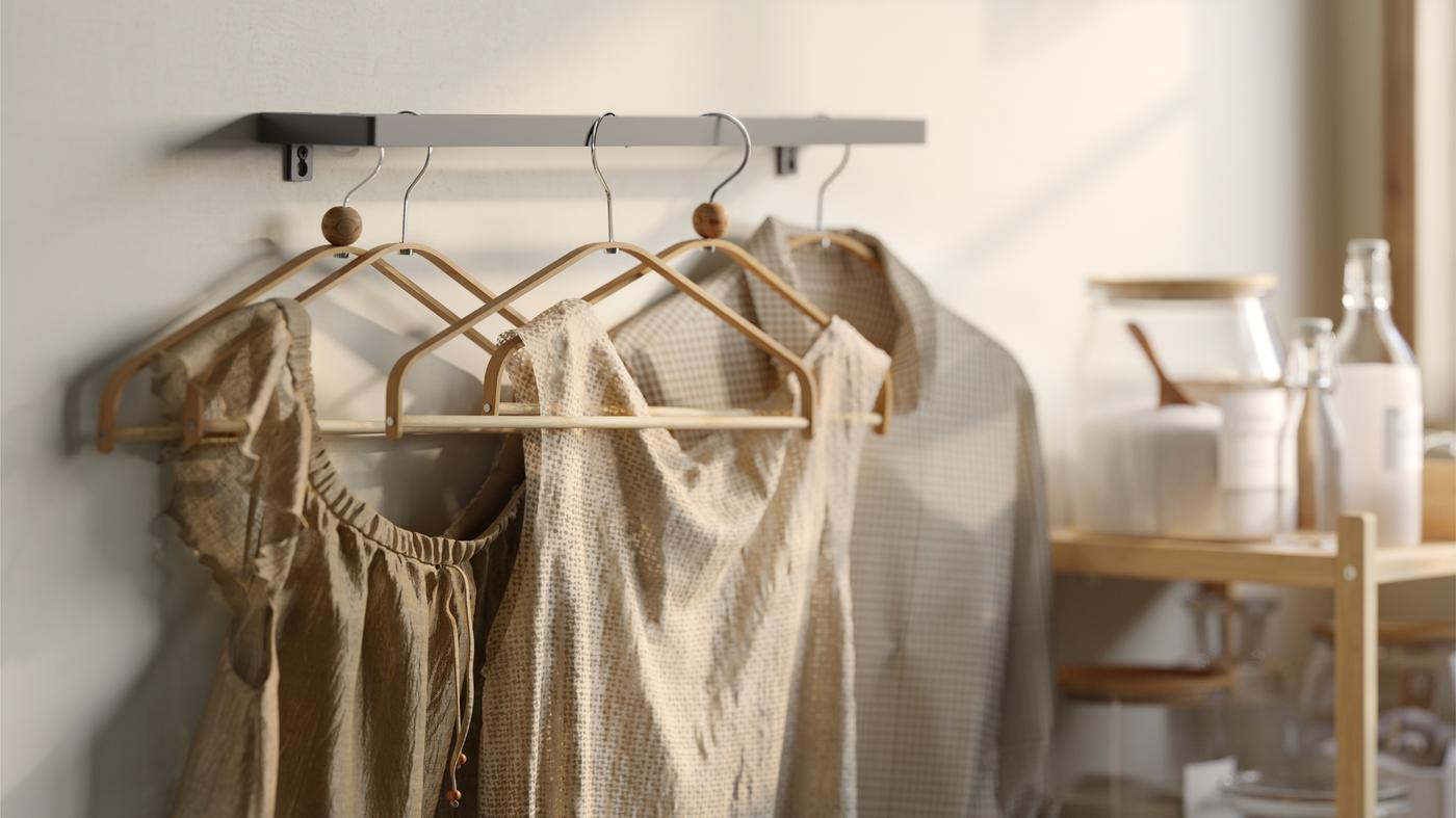Diverse grucce HÖSVANS in bambù su un binario in metallo montato a parete con camicie e vestiti beige accanto a un tavolo - IKEA.