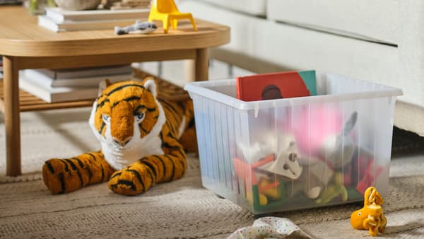DJUNGELSKOG tiger plush toy next to a VESSLA storage box with wheels in the living room with various toys stored inside. A BLAVINGAD octopus is also lying on the ground next to the box. 