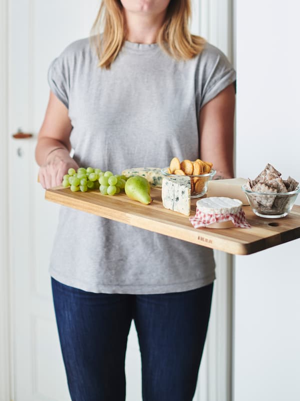 Donna in jeans e t-shirt che porta un tagliere SMÅÄTA con frutta, salatini e formaggi vari - IKEA