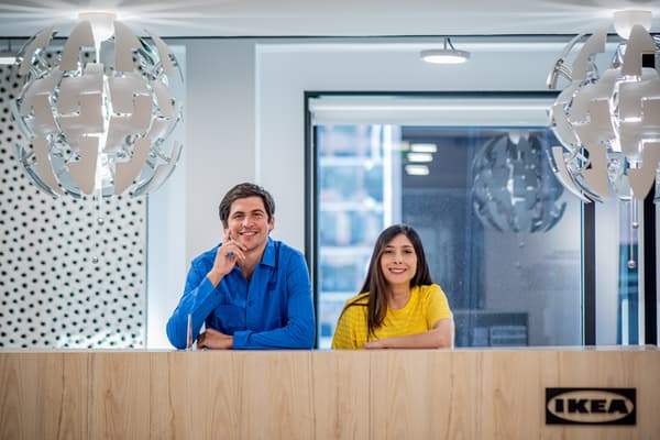 Dos colaboradores, en polera azul y amarilla, sonriendo. Se encuentran detrás del mesón de recepción de las oficinas IKEA.
