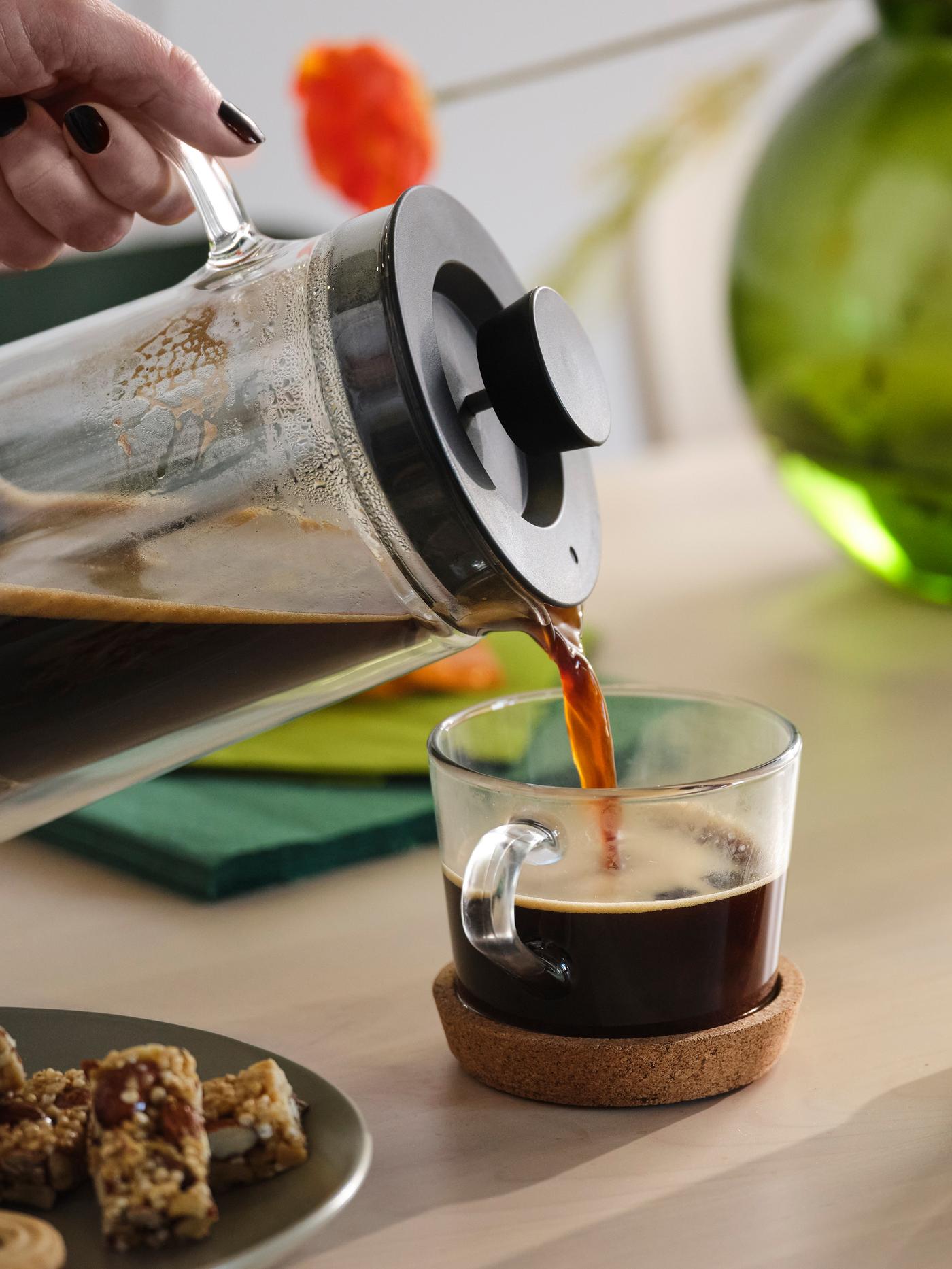 Du café est versé d’une cafetière dans une tasse posée sur un sous-verre en liège. À l’arrière-plan, un vase vert et une fleur orange.