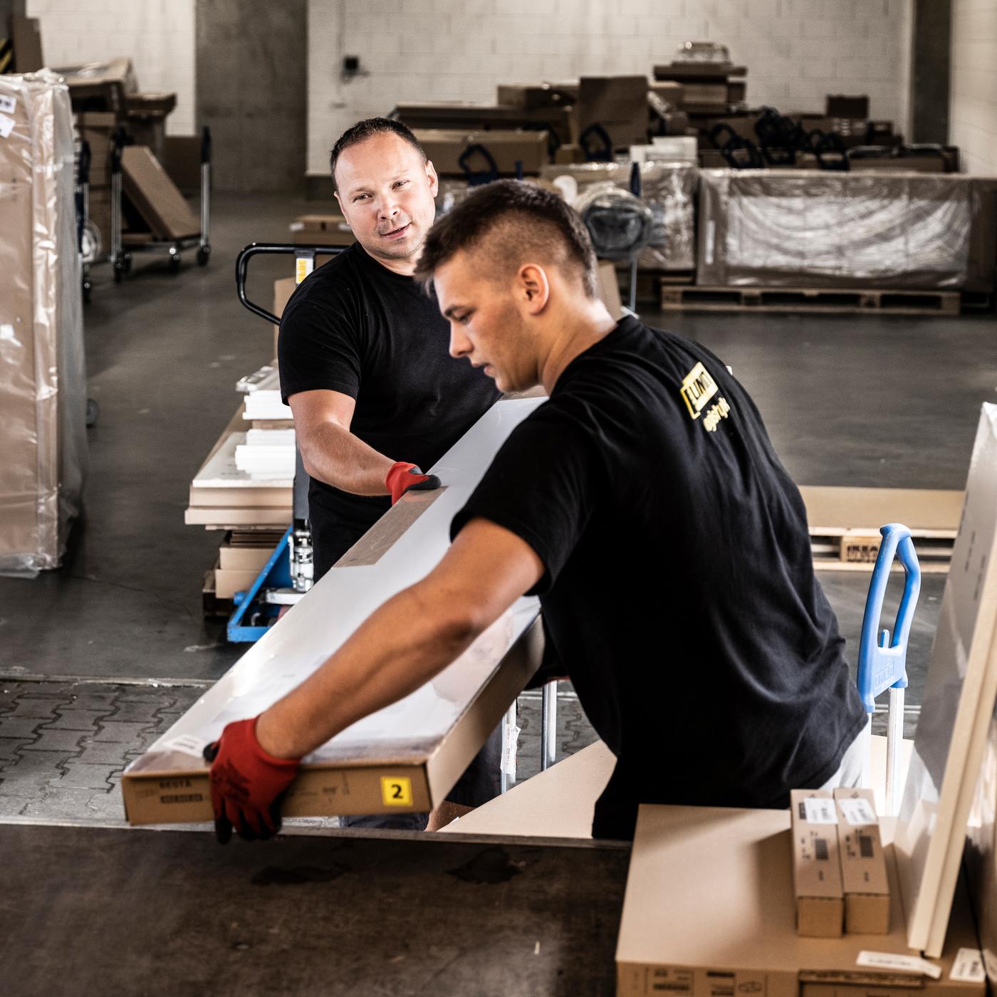Dua orang lelaki yang memakai kemeja-T berwarna hitam dan sarung tangan kerja berwarna merah sedang memunggah pek leper IKEA dari troli ke dalam bahagian belakang trak.