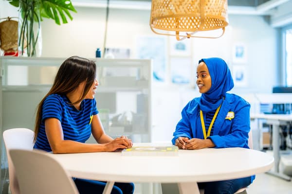Due dipendenti IKEA chiacchierano a un tavolo rotondo moderno in un'area di lavoro luminosa e contemporanea, circondati da piante e luce naturale.